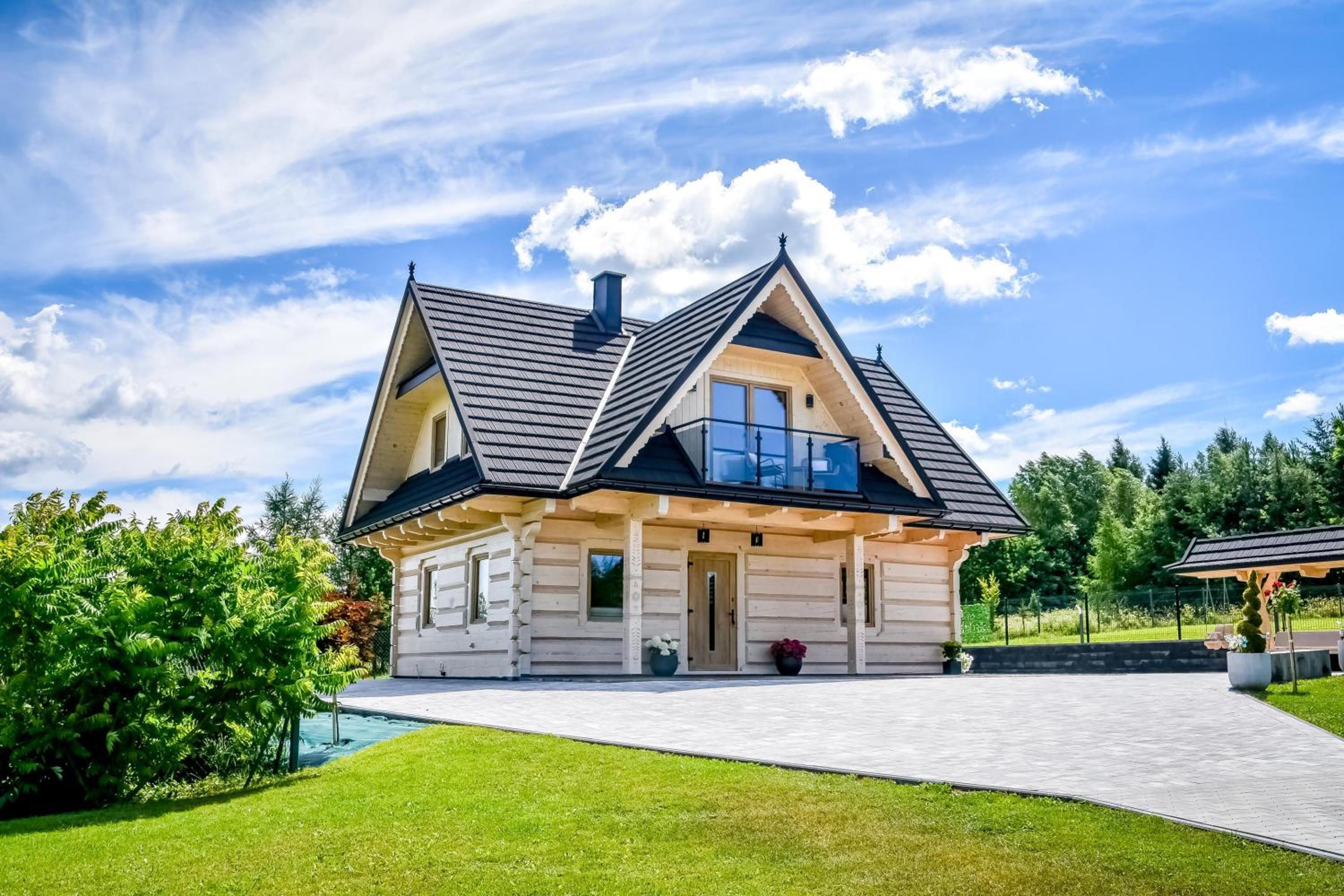 Domek z bali z ciemnym dachem i balkonem, przed nim trawnik, nad nim błękitne niebo
